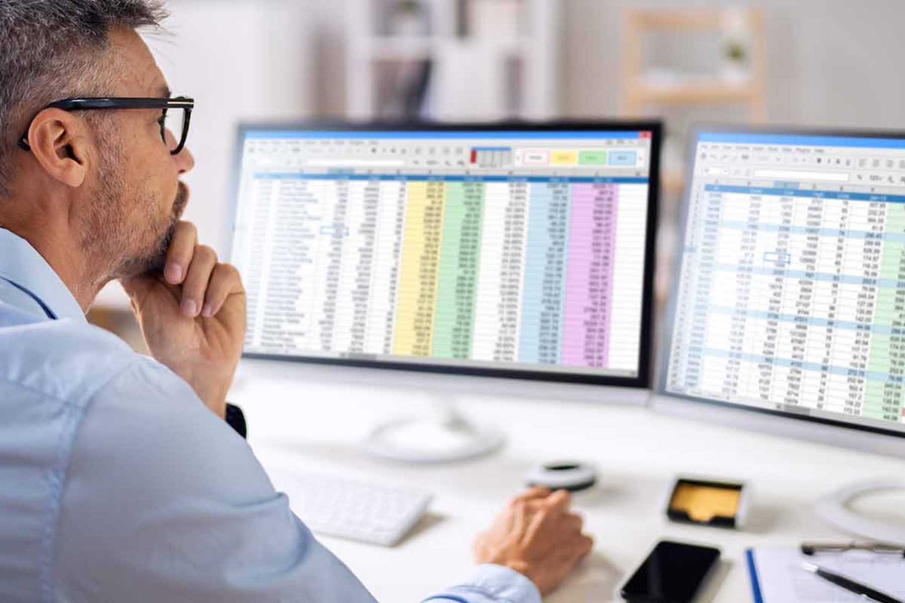 A man intently reviewing spreadsheets on dual computer screens, engaged in detailed data examination.